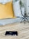 A small black book titled 'New Testament NIV' lies on a wooden surface. In the background, there is a cozy setup with a yellow pillow, a striped pillow, and some decorative plants. An abstract black geometric sculpture is also visible.