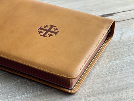 A close-up of a richly textured leather-bound book with gold embossed lettering on its spine, resting on a wooden table bathed in warm sunlight.