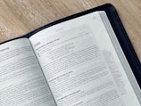 An open book displaying a page from the Gospel of Mark in the New Testament. The text is printed in black on white paper, and the book is bound in a black cover. The book is placed on a light-colored wooden surface, and the page appears to be from a modern Bible.