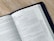 An open book displaying a page from the Gospel of Mark in the New Testament. The text is printed in black on white paper, and the book is bound in a black cover. The book is placed on a light-colored wooden surface, and the page appears to be from a modern Bible.
