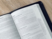 An open book displaying a page from the Gospel of Mark in the New Testament. The text is printed in black on white paper, and the book is bound in a black cover. The book is placed on a light-colored wooden surface, and the page appears to be from a modern Bible.