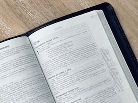 An open book displaying a page from the Gospel of Mark in the New Testament. The text is printed in black on white paper, and the book is bound in a black cover. The book is placed on a light-colored wooden surface, and the page appears to be from a modern Bible.