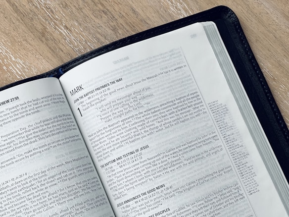 An open book displaying a page from the Gospel of Mark in the New Testament. The text is printed in black on white paper, and the book is bound in a black cover. The book is placed on a light-colored wooden surface, and the page appears to be from a modern Bible.