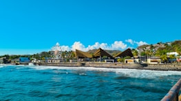 A coastal amusement area featuring a large ferris wheel and triangular-roofed structures. The blue ocean waves crash against a stone barrier, with hills and lush vegetation in the background. Bright sun and a clear sky enhance the vibrant, tropical atmosphere.