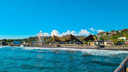 A coastal amusement area featuring a large ferris wheel and triangular-roofed structures. The blue ocean waves crash against a stone barrier, with hills and lush vegetation in the background. Bright sun and a clear sky enhance the vibrant, tropical atmosphere.