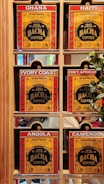 A display of six coffee boxes labeled with different countries: Ghana, Haiti, Ivory Coast, Central African Republic (abbreviated as Cent. African), Angola, and Cameroon. The boxes are primarily red and yellow with the brand name 'Bacha Coffee' prominently displayed. Each box is positioned within a wooden shelf, and there are some greenery and reflections visible in the background.