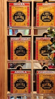 A display of six coffee boxes labeled with different countries: Ghana, Haiti, Ivory Coast, Central African Republic (abbreviated as Cent. African), Angola, and Cameroon. The boxes are primarily red and yellow with the brand name 'Bacha Coffee' prominently displayed. Each box is positioned within a wooden shelf, and there are some greenery and reflections visible in the background.