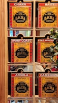 A display of six coffee boxes labeled with different countries: Ghana, Haiti, Ivory Coast, Central African Republic (abbreviated as Cent. African), Angola, and Cameroon. The boxes are primarily red and yellow with the brand name 'Bacha Coffee' prominently displayed. Each box is positioned within a wooden shelf, and there are some greenery and reflections visible in the background.