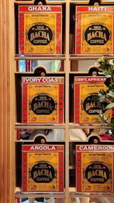 A display of six coffee boxes labeled with different countries: Ghana, Haiti, Ivory Coast, Central African Republic (abbreviated as Cent. African), Angola, and Cameroon. The boxes are primarily red and yellow with the brand name 'Bacha Coffee' prominently displayed. Each box is positioned within a wooden shelf, and there are some greenery and reflections visible in the background.