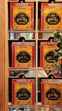 A display of six coffee boxes labeled with different countries: Ghana, Haiti, Ivory Coast, Central African Republic (abbreviated as Cent. African), Angola, and Cameroon. The boxes are primarily red and yellow with the brand name 'Bacha Coffee' prominently displayed. Each box is positioned within a wooden shelf, and there are some greenery and reflections visible in the background.