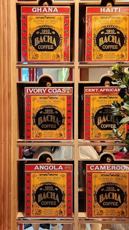 A display of six coffee boxes labeled with different countries: Ghana, Haiti, Ivory Coast, Central African Republic (abbreviated as Cent. African), Angola, and Cameroon. The boxes are primarily red and yellow with the brand name 'Bacha Coffee' prominently displayed. Each box is positioned within a wooden shelf, and there are some greenery and reflections visible in the background.