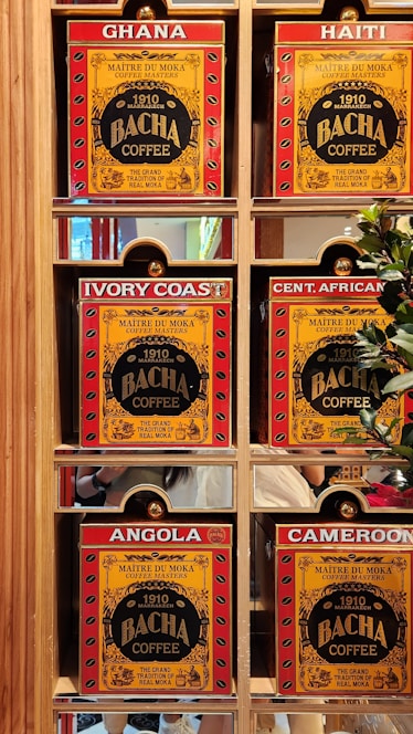 A display of six coffee boxes labeled with different countries: Ghana, Haiti, Ivory Coast, Central African Republic (abbreviated as Cent. African), Angola, and Cameroon. The boxes are primarily red and yellow with the brand name 'Bacha Coffee' prominently displayed. Each box is positioned within a wooden shelf, and there are some greenery and reflections visible in the background.