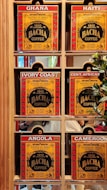 A display of six coffee boxes labeled with different countries: Ghana, Haiti, Ivory Coast, Central African Republic (abbreviated as Cent. African), Angola, and Cameroon. The boxes are primarily red and yellow with the brand name 'Bacha Coffee' prominently displayed. Each box is positioned within a wooden shelf, and there are some greenery and reflections visible in the background.