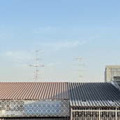 A roof with a corrugated metal surface and a geometric metal railing in front. Multiple television antennas extend upwards against a clear blue sky. Part of a tall building is visible in the background.
