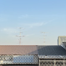A roof with a corrugated metal surface and a geometric metal railing in front. Multiple television antennas extend upwards against a clear blue sky. Part of a tall building is visible in the background.