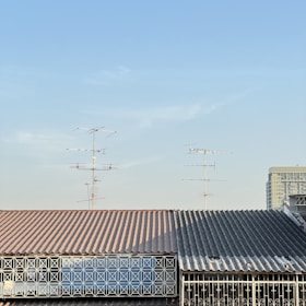 A roof with a corrugated metal surface and a geometric metal railing in front. Multiple television antennas extend upwards against a clear blue sky. Part of a tall building is visible in the background.