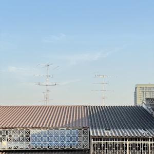 A roof with a corrugated metal surface and a geometric metal railing in front. Multiple television antennas extend upwards against a clear blue sky. Part of a tall building is visible in the background.