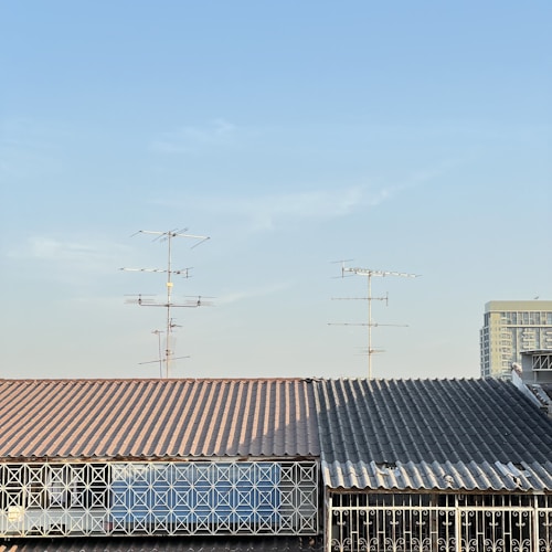 A roof with a corrugated metal surface and a geometric metal railing in front. Multiple television antennas extend upwards against a clear blue sky. Part of a tall building is visible in the background.