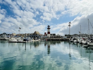 A picturesque marina with several boats docked along the wooden piers. In the background, a prominent red and white lighthouse stands tall. The marina is surrounded by quaint buildings, and there are a few tall palm trees. The water is calm and reflects the cloudy sky above.