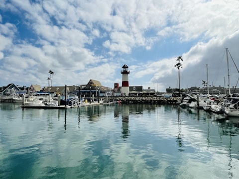 A picturesque marina with several boats docked along the wooden piers. In the background, a prominent red and white lighthouse stands tall. The marina is surrounded by quaint buildings, and there are a few tall palm trees. The water is calm and reflects the cloudy sky above.