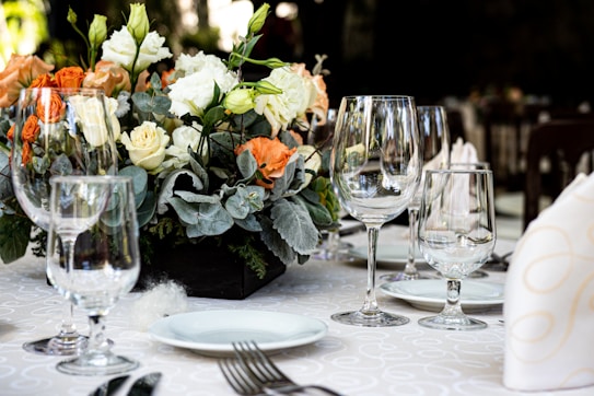 A beautifully arranged dining table featuring an elegant floral centerpiece with orange, cream, and white flowers. Surrounding the centerpiece are several clear wine glasses and plates, neatly set with napkins and silverware atop a white tablecloth with a subtle pattern.