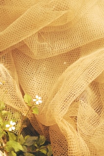 A textured, light burlap or mesh fabric forms soft folds, creating a rustic and organic aesthetic. In the foreground, small white flowers with green leaves delicately emerge, contrasting with the fabric's earthy tones.