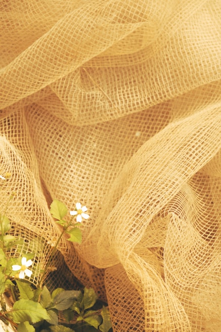 A textured, light burlap or mesh fabric forms soft folds, creating a rustic and organic aesthetic. In the foreground, small white flowers with green leaves delicately emerge, contrasting with the fabric's earthy tones.