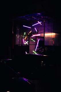 A dimly lit scene featuring a storefront with vibrant, neon lights. The glass facade is adorned with various colorful, glowing rods arranged in a dynamic pattern. The neon lights include shades of purple, pink, green, and blue. The name of the establishment is partially visible in script, adding a sense of mystery and intrigue to the setting. The ambiance is moody and somewhat industrial.