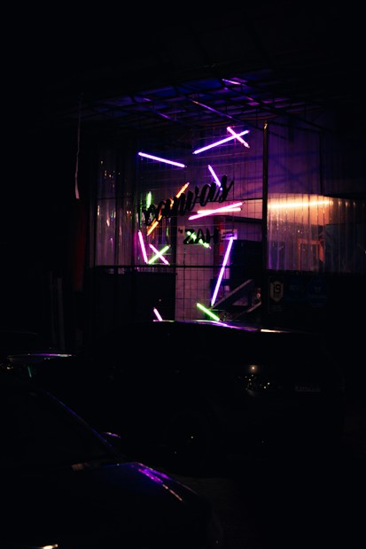 A dimly lit scene featuring a storefront with vibrant, neon lights. The glass facade is adorned with various colorful, glowing rods arranged in a dynamic pattern. The neon lights include shades of purple, pink, green, and blue. The name of the establishment is partially visible in script, adding a sense of mystery and intrigue to the setting. The ambiance is moody and somewhat industrial.