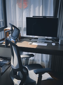 A modern home office setup with a sleek desk featuring a large computer monitor, wireless keyboard, and mouse. A comfortable office chair is situated in front of the desk. Books are stacked neatly on the left side. Sunlight streams in through sheer curtains, casting soft shadows across the workspace.