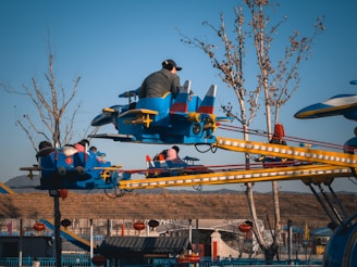 A vibrant amusement park ride featuring blue and yellow airplane-shaped cars suspended on metal arms, set against a clear blue sky. The ride is occupied by several people, including children and an adult wearing a cap, experiencing enjoyment as the ride spins them around. Bare trees with sparse branches are visible in the background along with some decorative elements like red lanterns.