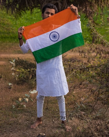 A person in a white traditional outfit stands outdoors, holding an Indian flag with an orange, white, and green tricolor and a blue Ashoka Chakra in the center. The background consists of green foliage and brownish grass, indicating a natural setting.