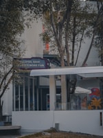 An outdoor setting featuring a modern building facade, partially obscured by trees. Visible signage reads 'Alcohol Bank,' suggesting a fun or leisure-oriented establishment. The area is surrounded by vegetation, with glass windows and doors visible, contributing to a calm and inviting atmosphere.
