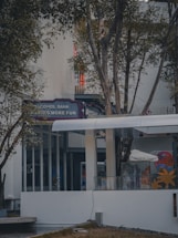 An outdoor setting featuring a modern building facade, partially obscured by trees. Visible signage reads 'Alcohol Bank,' suggesting a fun or leisure-oriented establishment. The area is surrounded by vegetation, with glass windows and doors visible, contributing to a calm and inviting atmosphere.