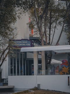 An outdoor setting featuring a modern building facade, partially obscured by trees. Visible signage reads 'Alcohol Bank,' suggesting a fun or leisure-oriented establishment. The area is surrounded by vegetation, with glass windows and doors visible, contributing to a calm and inviting atmosphere.