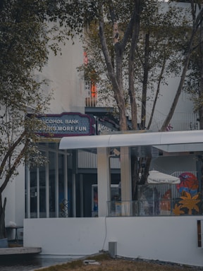 An outdoor setting featuring a modern building facade, partially obscured by trees. Visible signage reads 'Alcohol Bank,' suggesting a fun or leisure-oriented establishment. The area is surrounded by vegetation, with glass windows and doors visible, contributing to a calm and inviting atmosphere.