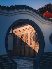 A traditional Chinese architectural element is visible through a round moon gate, featuring elaborately carved wooden panels illuminated by sunlight. A red lantern hangs above, adding a touch of vibrancy to the scene.
