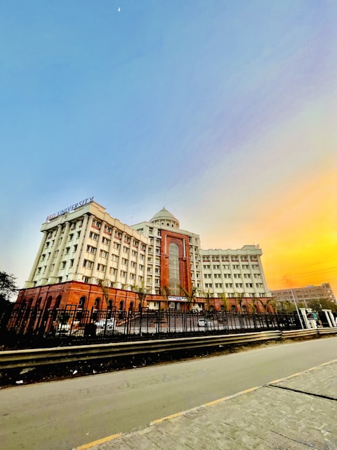 A large multi-story building with a grand entrance featuring prominent arches and windows. The structure is surrounded by a black fence and is situated alongside a wide road. The building has a dome at the top and displays the name 'BBD University'. The sky is clear with a gradient of light blue transitioning into orange hues as if during sunrise or sunset, with the moon faintly visible.