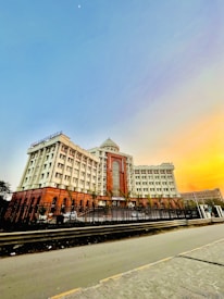 A large multi-story building with a grand entrance featuring prominent arches and windows. The structure is surrounded by a black fence and is situated alongside a wide road. The building has a dome at the top and displays the name 'BBD University'. The sky is clear with a gradient of light blue transitioning into orange hues as if during sunrise or sunset, with the moon faintly visible.