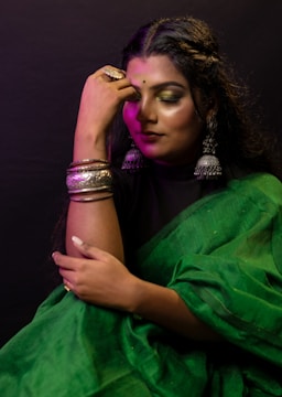 A woman sits wearing a vibrant green saree and silver jewelry, including large earrings and bangles. Her makeup is dramatic with golden eyeshadow and bold lipstick. The lighting casts a soft, warm glow, highlighting her serene expression.
