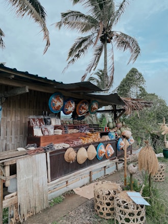 A rustic outdoor market stall made of bamboo, displaying an assortment of traditional handwoven hats and decorative items. Various packaged goods and local foods are arranged on the counter. Large painted circular items hang overhead as decoration. The setting includes large palm trees swaying in the breeze, contributing to a tropical vibe.