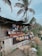 A rustic outdoor market stall made of bamboo, displaying an assortment of traditional handwoven hats and decorative items. Various packaged goods and local foods are arranged on the counter. Large painted circular items hang overhead as decoration. The setting includes large palm trees swaying in the breeze, contributing to a tropical vibe.