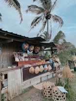 A rustic outdoor market stall made of bamboo, displaying an assortment of traditional handwoven hats and decorative items. Various packaged goods and local foods are arranged on the counter. Large painted circular items hang overhead as decoration. The setting includes large palm trees swaying in the breeze, contributing to a tropical vibe.