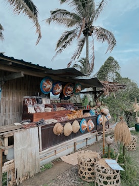 A rustic outdoor market stall made of bamboo, displaying an assortment of traditional handwoven hats and decorative items. Various packaged goods and local foods are arranged on the counter. Large painted circular items hang overhead as decoration. The setting includes large palm trees swaying in the breeze, contributing to a tropical vibe.