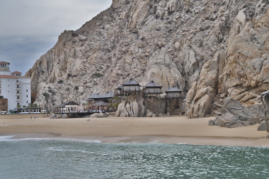 A coastal scene featuring a large rocky cliff with several structures nestled between the rocks. In the foreground, there is a sandy beach and a small wave breaking on the shore. To the left, a tall white and brown building stands close to the water, suggesting a resort or hotel.