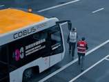 An airport shuttle bus labeled COBUS 3000 is parked on a tarmac area with two individuals wearing uniforms walking near it. The bus has visible branding for Shenzhen Baoan International Airport and features an orange roof with a small beacon light.
