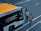 An airport shuttle bus labeled COBUS 3000 is parked on a tarmac area with two individuals wearing uniforms walking near it. The bus has visible branding for Shenzhen Baoan International Airport and features an orange roof with a small beacon light.