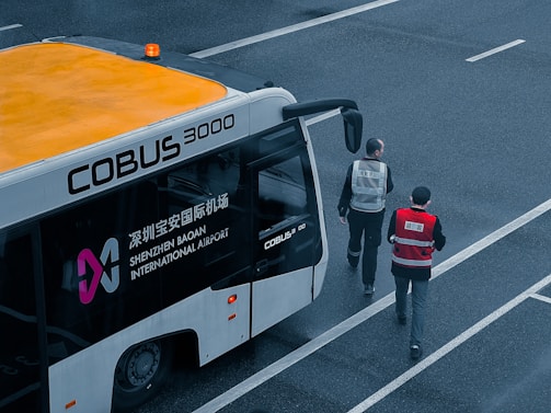 An airport shuttle bus labeled COBUS 3000 is parked on a tarmac area with two individuals wearing uniforms walking near it. The bus has visible branding for Shenzhen Baoan International Airport and features an orange roof with a small beacon light.