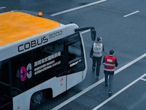 An airport shuttle bus labeled COBUS 3000 is parked on a tarmac area with two individuals wearing uniforms walking near it. The bus has visible branding for Shenzhen Baoan International Airport and features an orange roof with a small beacon light.