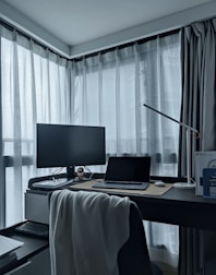 A modern home office setup with a sleek desk positioned by large windows with sheer curtains. The desktop surface includes a monitor, a closed laptop, a desk lamp, and a small figurine. A chair with a blanket draped over the back is in front of the desk, and the overall setting appears organized and minimalist.